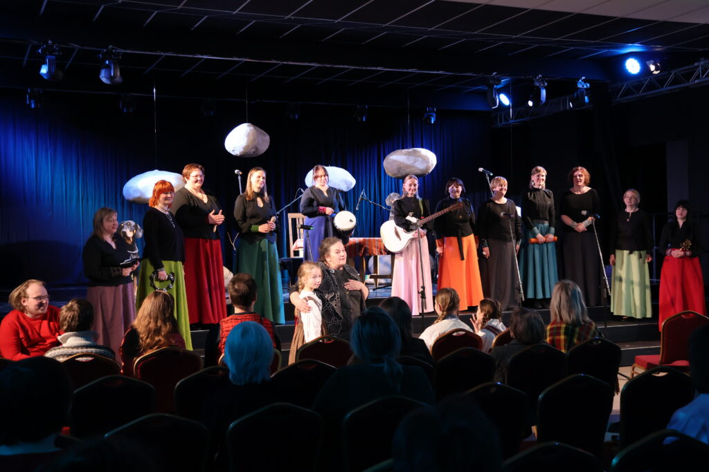 Stāstnieku festivāla Ziv zup koncertā Cik augšā sīku zvaigžņu, zemē sīku akmentiņu dalībnieki vienojas tautasdziesmā Upe nesa ozoliņu. Uz skatuves – Padures ansamblis Harmonija, centrā festivāla rīkotāja Ina Celitāne un mazā stāstniece Gerda Gruze.